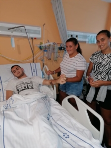 Christian in a hospital bed holding hands with Tracy, Carmen stands next to her.